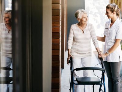 Pflegerin führt ältere Frau mit Rollator über den Hof